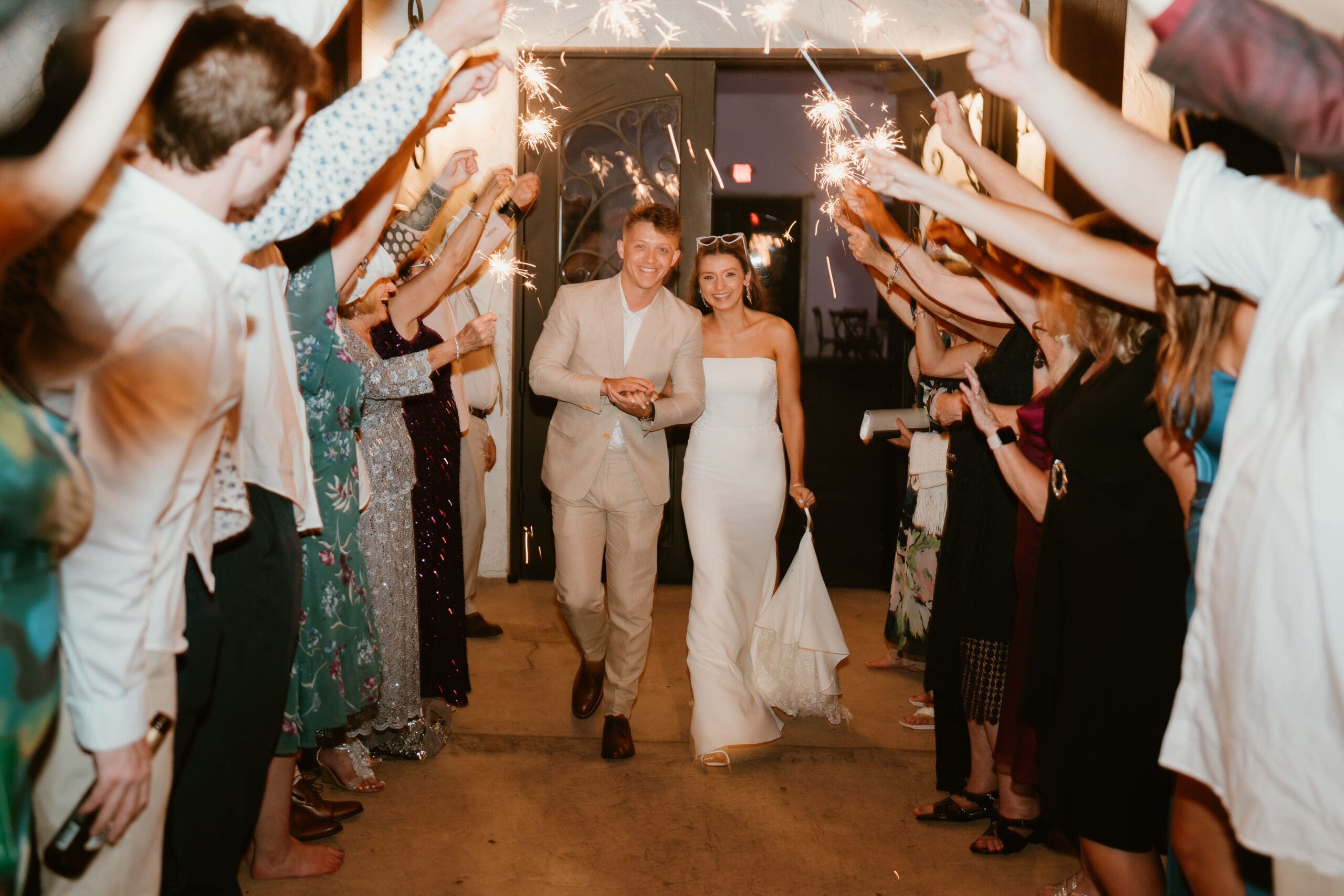 Bride and groom walking through a sparkler tunnel during wedding sendoff, surrounded by cheering guests in an evening celebration